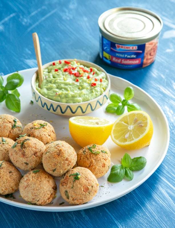 baked salmon meatballs on white plate served with halved lemon, basil leaves and avocado cream in a small pot, can of salmon in the background