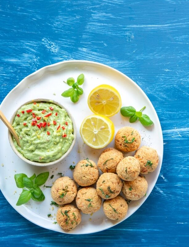 flat lay image of baked salmon meatballs on white plate served with halved lemon, basil leaves and avocado cream in a small pot