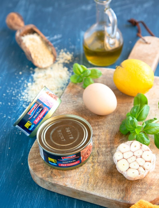 baked salmon meatballs ingredients: canned salmon, garlic, basil, lemon and egg on a wood board, almond flour and olive oil next to it