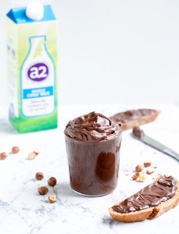Italian chocolate hazelnut spread in large clear glass, bread slice with spread on the right side, knife with spread, a2 milk carton and hazelnuts on the left side