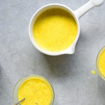 Golden moon milk in small white saucepan next to three mugs filled with golden moon milk on grey background