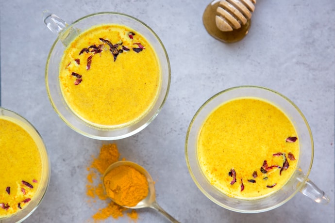 Golden moon milk in three mugs topped with hibiscus flowers, next to teaspoon with ground turmeric and honey drizzler with honey, on grey background