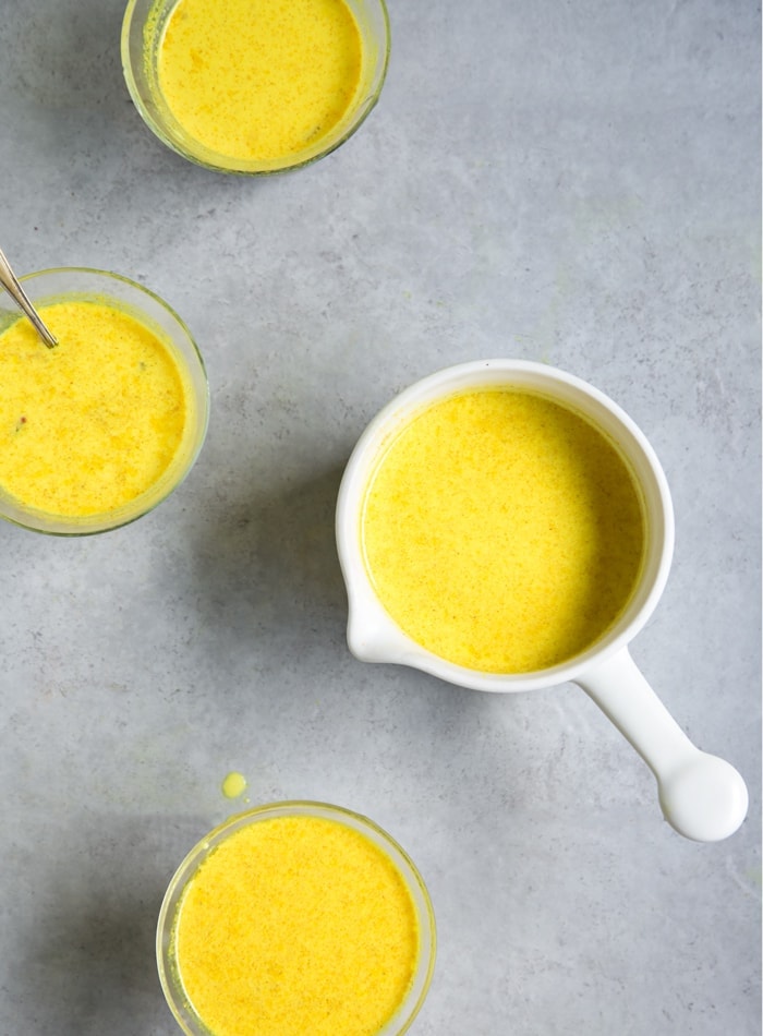 Golden moon milk in small white saucepan next to three mugs filled with golden moon milk on grey background