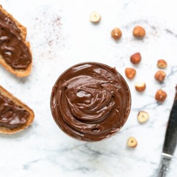 Italian chocolate hazelnut spread flat lay image featuring glass full of spread, and bread slices with spread both on the top right and on the bottom left, hazelnuts spread around for decoration and knife with spread on the bottom right