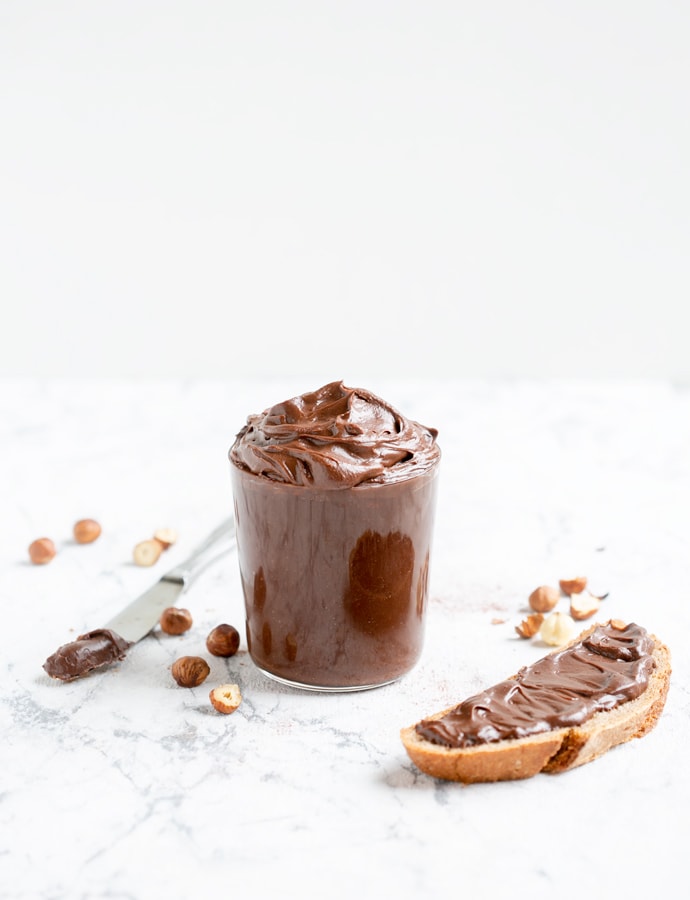 Italian chocolate hazelnut spread in large clear glass, bread slice with spread on the right side, knife with spread and hazelnuts on the left side