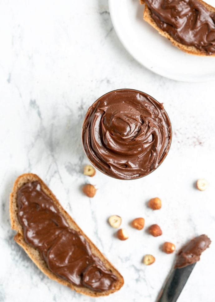Italian chocolate hazelnut spread flat lay image featuring glass full of spread, and bread slices with spread both on the top right and on the bottom left, hazelnuts spread around for decoration and knife with spread on the bottom right
