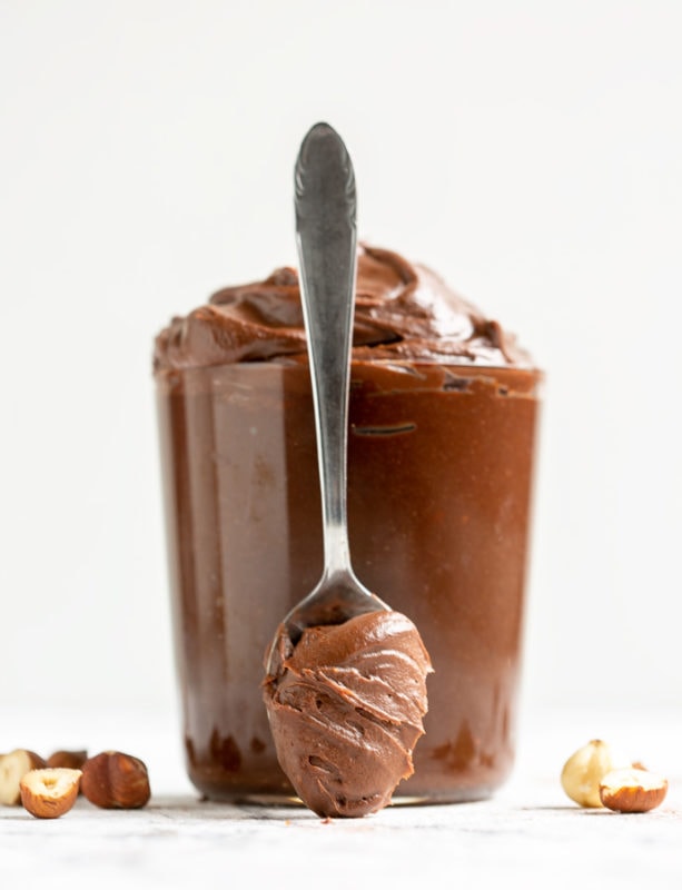 Italian chocolate hazelnut spread in large clear glass and spoon with chocolate hazelnut spread in front of it, hazelnuts on the sides for decoration