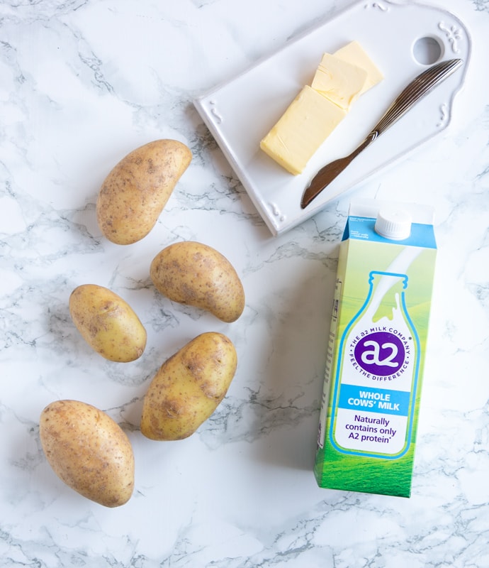 mashed potato recipe ingredients: five potatoes, butter and butter knife on white cutting board and carton of a2 Milk