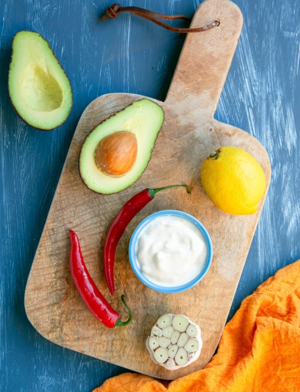 avocado dip ingredients: halved avocado, lemon, pot with yogurt, 2 red chili, halved garlic on a wood board, orange napkin next to the board