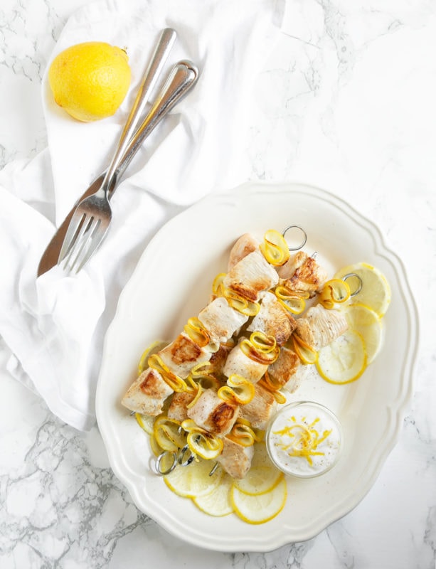 flat-lay image of lemon chicken skewers and small pot with yogurt dip and lemon slices onto white serving plate. Lemon and for on a white napkin in the backgroun