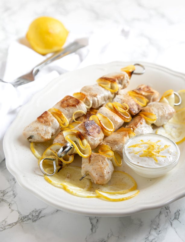 lemon chicken skewers and small pot with yogurt dip and lemon slices onto white serving plate. Lemon and for on a white napkin in the backgroun