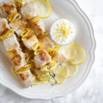 lemon chicken skewers and small pot with yogurt dip and lemon slices onto white serving plate