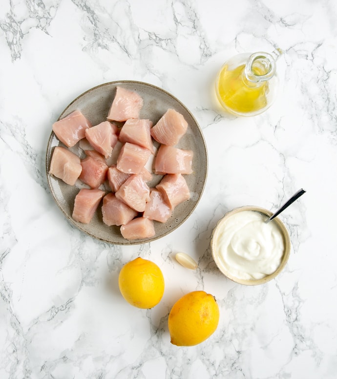 chicken cubes onto a grey plate, two lemons, one garlic clove, yogurt in a small pot with a teaspoon and olive oil in a glass bottle