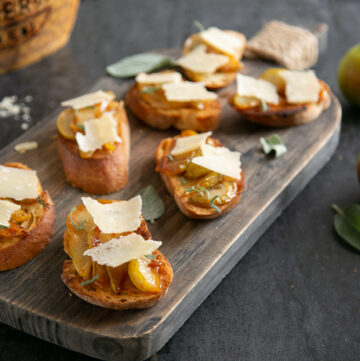 Bruschetta with Caramelised Pears & Grana Padano Riserva on a wood board, in the background wheel of grana padano cheese, cheese knife and two pears.