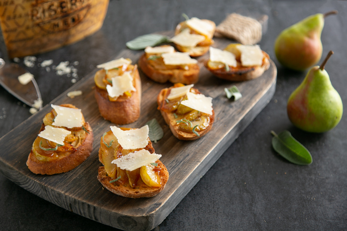 Bruschetta with Caramelised Pears & Grana Padano Riserva on a wood board, in the background wheel of grana padano cheese, cheese knife and two pears.