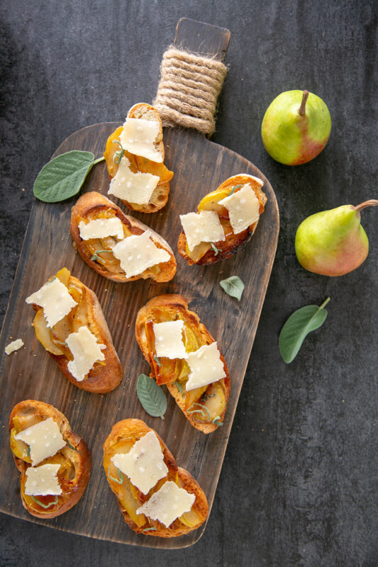 Bruschetta with Caramelised Pears and Grana Padano Riserva on a wood board, scattered sage leaves around and two pears on the top left side