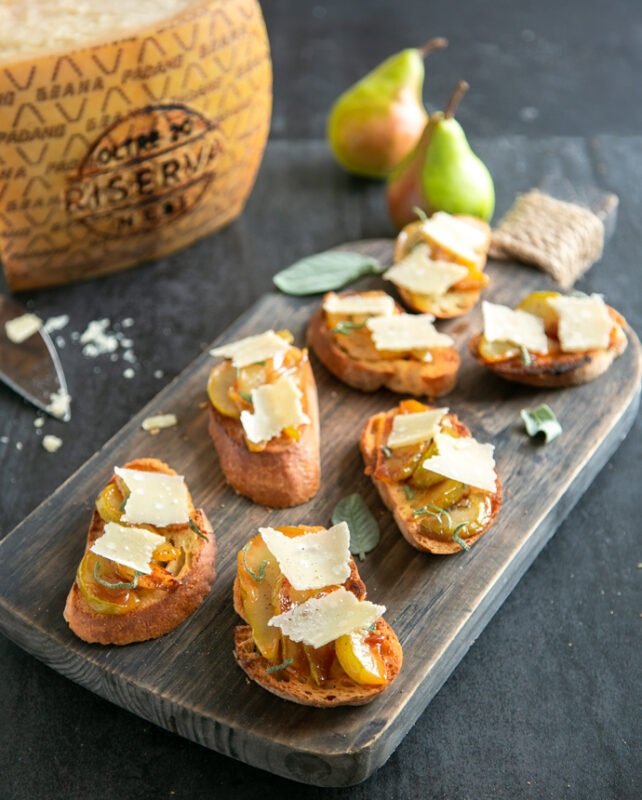 Bruschetta with Caramelised Pears & Grana Padano Riserva on a wood board, in the background wheel of grana padano cheese, cheese knife and two pears.