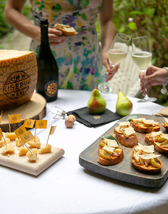 Table with a woodboard with Grana padano cheese pieces on the left, woodboard with bruschetta with caramelised pears on the right, 2 pears on the back, lady holding a bruschetta in one hand and a glass of prosecco into the other toasting with another glass of prosecco. Prosecco bottle and wheel of grana padano cheese next to the lady.