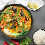 Thai Red Fish Curry in cast iron skillet, next to a white cloth with fresh ginger root, cilantro, red chili peppers and halved limes, and a small bowl of rice.