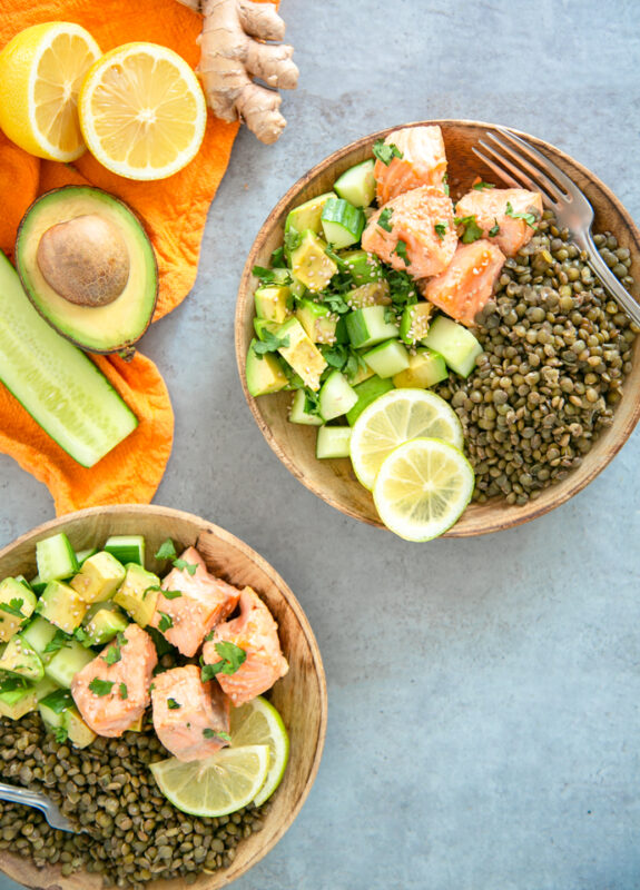 on the top left orange napkin with half cucumber, half avocado, halved lemon and ginger root on top. On the down left side, the same salmon, lentils and veggie dish on a wood plate