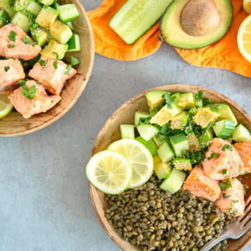 on the top right side an orange napkin with half cucumber, half avocado, halved lemon and ginger root on top. On the top left side, the same salmon, lentils and veggie dish on a wood plate