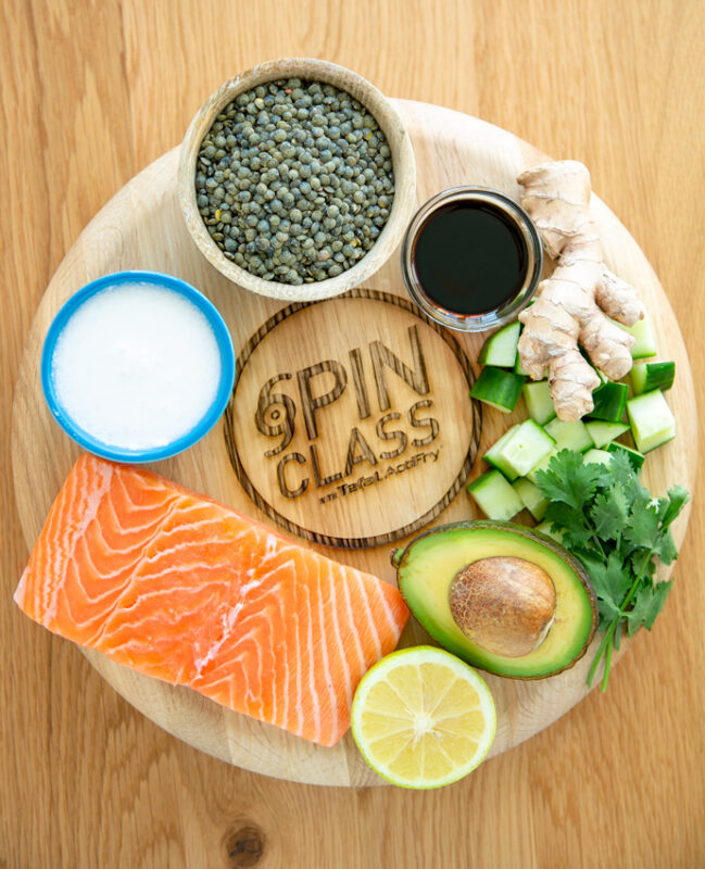 wood board branded with the Spin class by tefal actifry writing, on top of it a salmon fillet, small blue pot with coconut milk, small wood pot with green lentils, small glass pot with soy sauce, ginger root, cubed cucumber, coriander leaves, half avocado and half lemon.