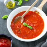 italian tomato sauce in a white pan with a wood spoon, next to a grey napkin on the right side, on the left side a small bowl with canned tomatoes, basil leaves and a small glass bottle of extra-virgin olive oil.