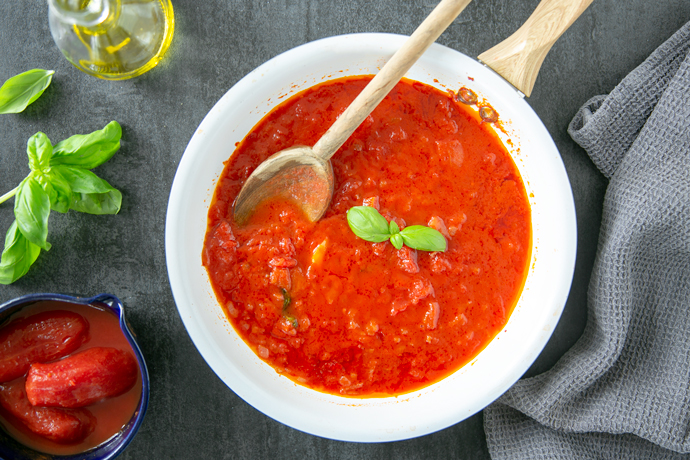 italian tomato sauce in a white pan, next to a grey napkin on the right side, on the left side a small bowl with canned tomatoes, basil leaves and a small glass bottle of extra-virgin olive oil.