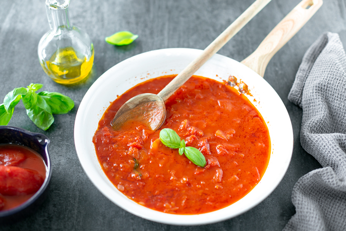 italian tomato sauce in a white pan with a wood spoon, next to a grey napkin on the right side, on the left side a small bowl with canned tomatoes, basil leaves and a small glass bottle of extra-virgin olive oil.
