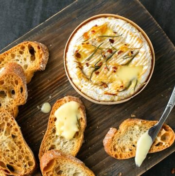 baked camembert in wooden box on a wooden board, next to a butter knife, toasted baguette slices, one bread slices with melted cheese on top, sprigs of rosemary and sea salt flakes.