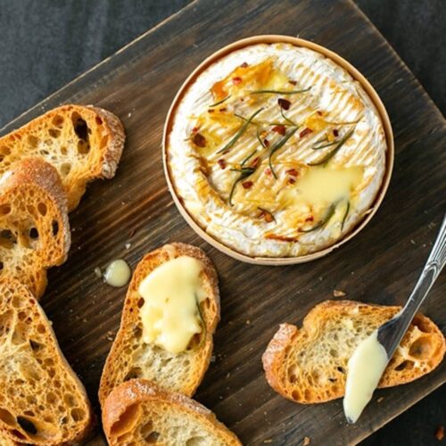 baked camembert in wooden box on a wooden board, next to a butter knife, toasted baguette slices, one bread slices with melted cheese on top, sprigs of rosemary and sea salt flakes.