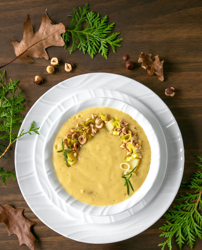 Leek and Potato soup in white bowl over two white plates, spoon on the side over a white napkin. brown and green leaves, and toasted hazelnuts scattered over the wood table