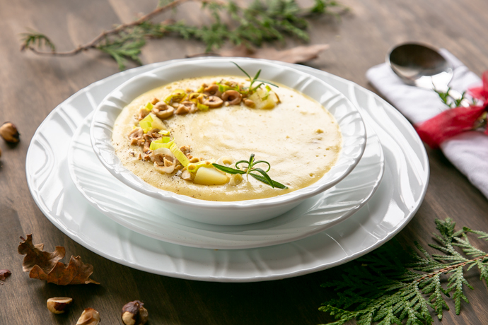 Leek and Potato soup in white bowl over two white plates, spoon on the side over a white napkin. brown and green leaves, and toasted hazelnuts scattered over the wood table