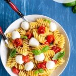 caprese pasta salad in a white bowl with serving spoons