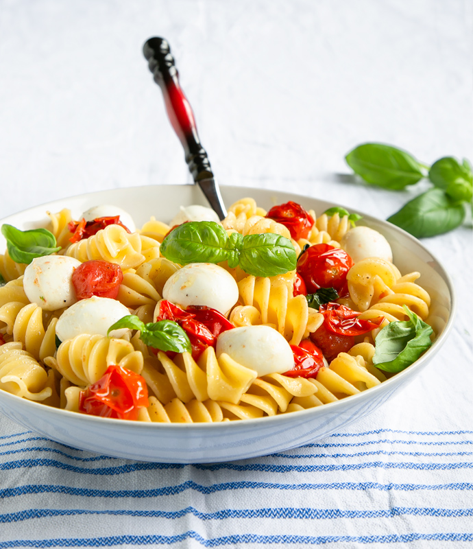Italian caprese pasta in a large white bowl with a serving spoon, basil leaves in the background