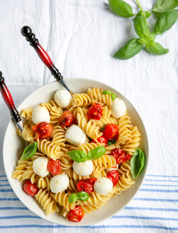 Italian caprese pasta in a large white bowl with serving spoons, basil leaves in the background