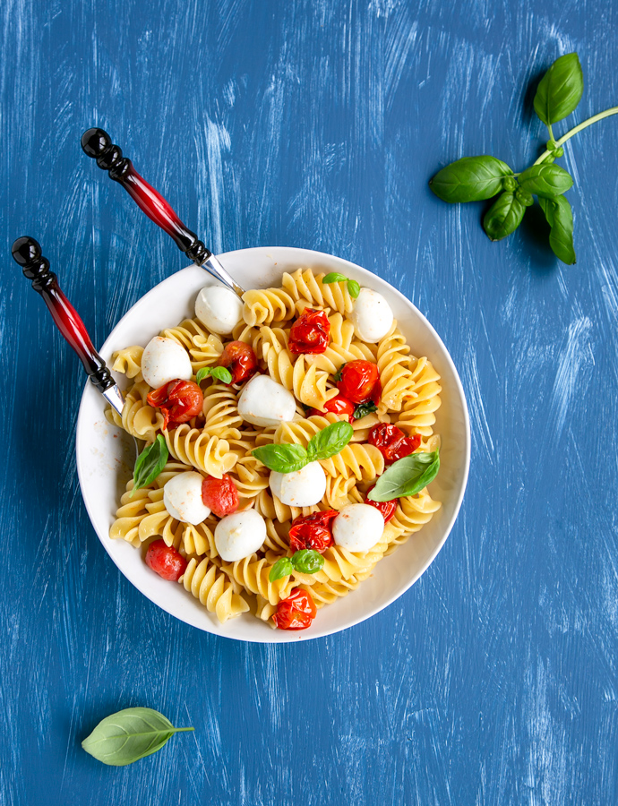 Italian caprese pasta in a large plate with serving spoons, basil leaves scattered over the blue background