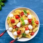 Italian caprese pasta salad in a large plate with serving spoons, basil leaves scattered over the blue background