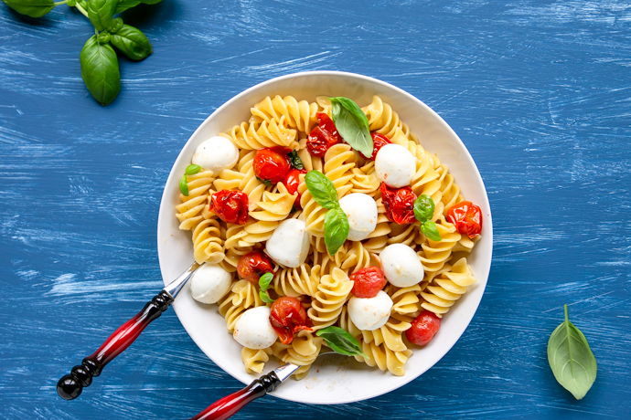 Italian caprese pasta salad in a large plate with serving spoons, basil leaves scattered over the blue background
