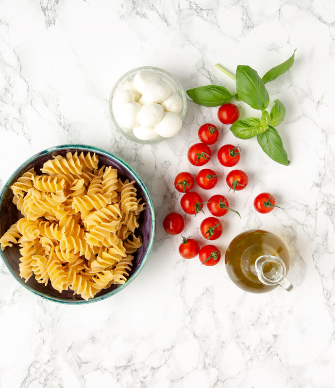 fusilli pasta in a large light blue pasta bowl, mozzarella balls in a small bowl, olive oil in glass bottle, basil leaves and cherry tomatoes scattered around the marble background
