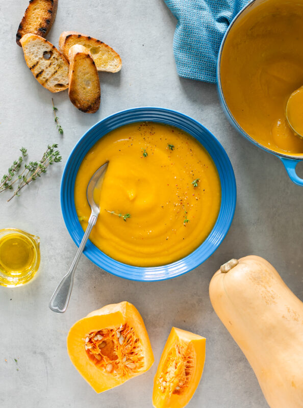 bowl of creamy butternut squash soup served with grilled bread, next to a pot with more soup, butternut squash slices,thyme sprigs, and small pot of olive oil