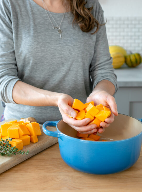 andrea holding chopped butternut squash over a cast iron pot, next to the lady butternut squash chunks over a wood board