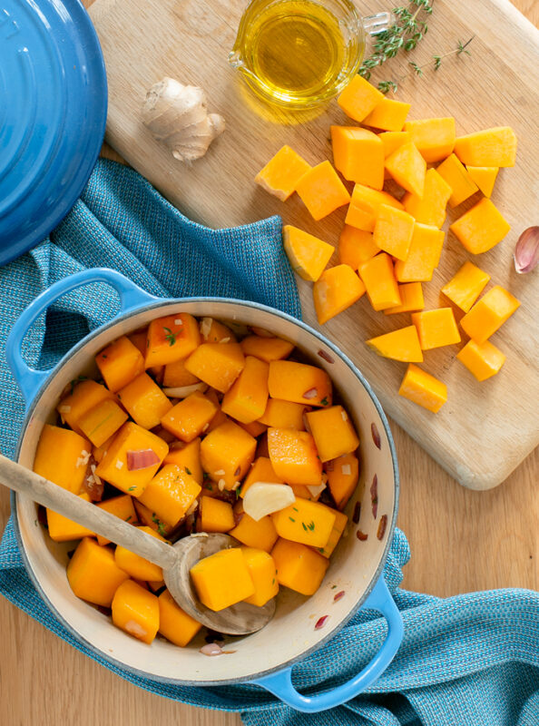 butternut squash soup recipe step 1: sauteed vegetables in a large cast iron pot, next to a wood board topped with butternut squash chunks, thyme sprigs, garlic, ginger piece and small just with olive oil