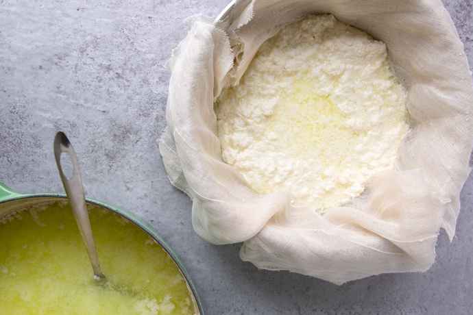 homemade ricotta over a bowl covered with cheese cloth, on the left side down part of the pot filled with the curds and a slotted spoon