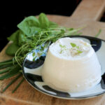 homemade ricotta over a small white an blue plate, next to fresh aromatic herbs, both over a wood board,