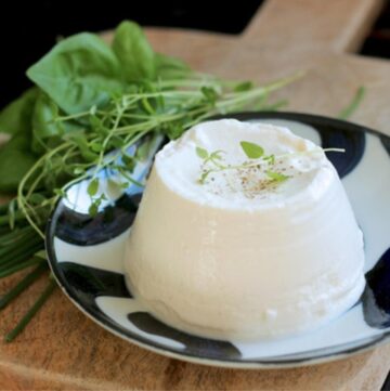 homemade ricotta over a small white an blue plate, next to fresh aromatic herbs, both over a wood board,