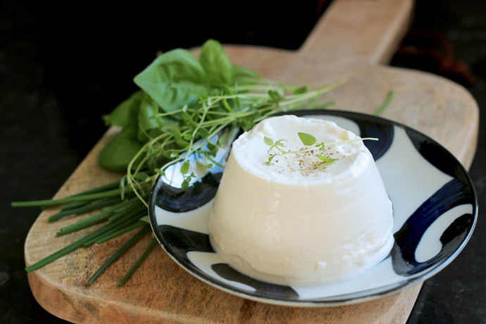 homemade ricotta over a small white an blue plate, next to fresh aromatic herbs, both over a wood board,