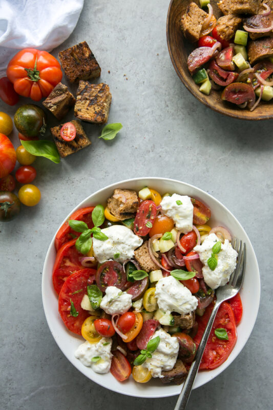 first recipe of 20-minute italian cookbook: burrata panzanella salad with a fork in a white place, tomatoes and grilled bread on the left side, small bowl with extra salad and bread on the right side