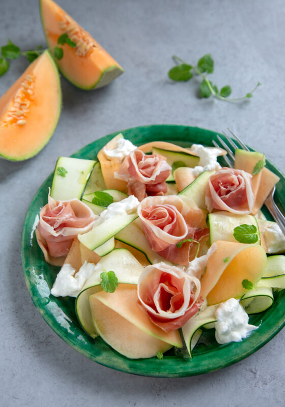 recipe from 20-minute italian cookbook: melon, zucchini, prosciutto and burrata salad on a green plate, two slices of melon in the background and fresh oragano leaves