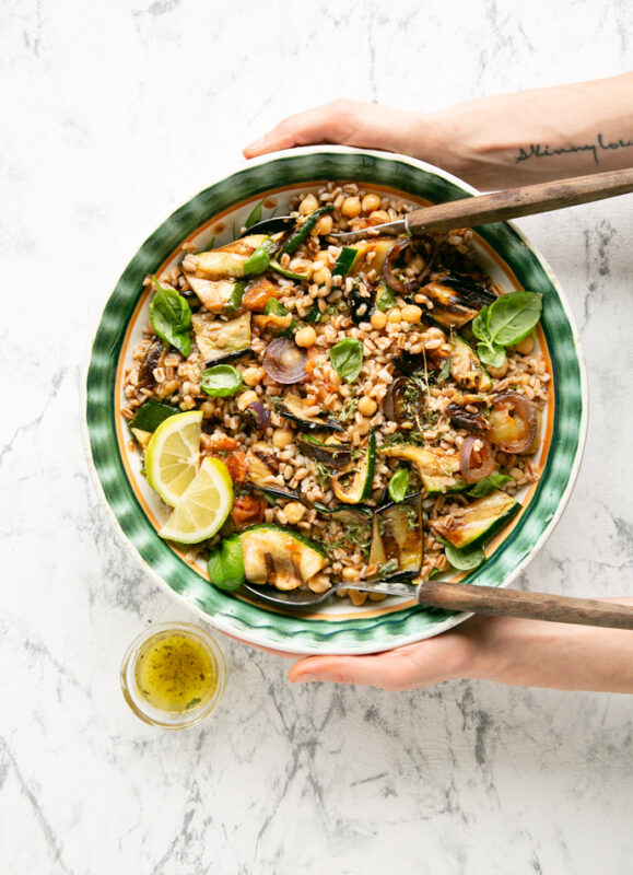 recipe from 20-minute italian cookbook: hands holding a bowl of farro salad with grilled vegetables, small pot of vinaigrette next to the salad